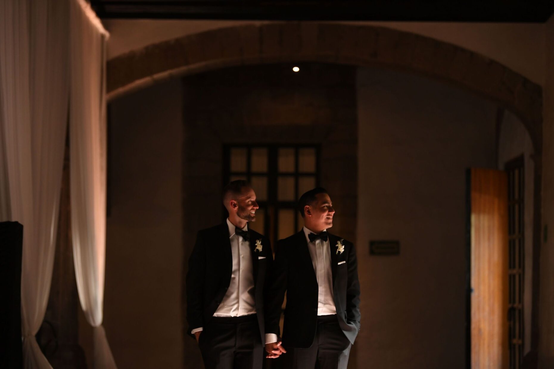 Two grooms holding hands in dim lighting wearing black tuxedos